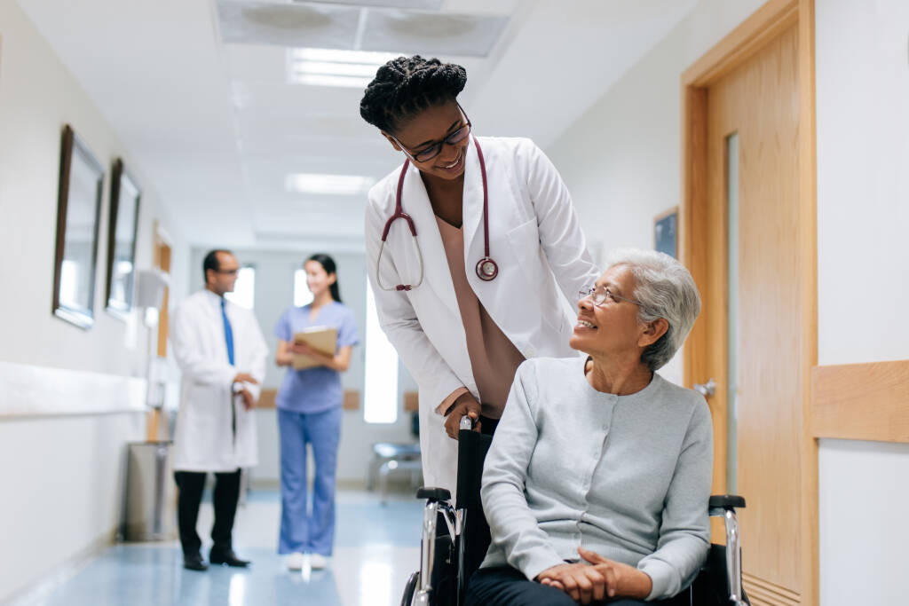 A female doctor pushing a senior patient on a wheelchair in a hospital, looking at each other and smiling.