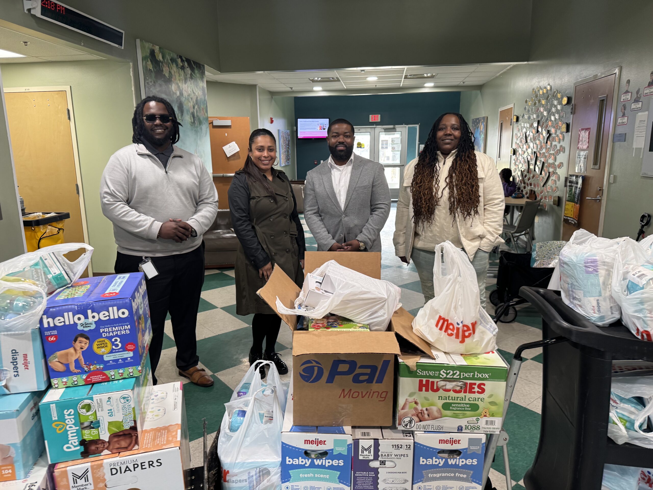 Four people pose with donated diapers, baby wipes, and infant care supplies collected during a Modivcare community donation drive for Alternatives for Girls.